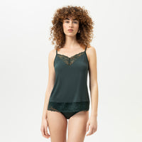 A model with curly brown hair wearing a dark green satin camisole and matching underwear with lace trim, photographed against a plain white studio background.