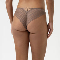 Rear view of a model wearing a taupe lace thong with a small gold clasp at the center back, set against a plain light gray background.