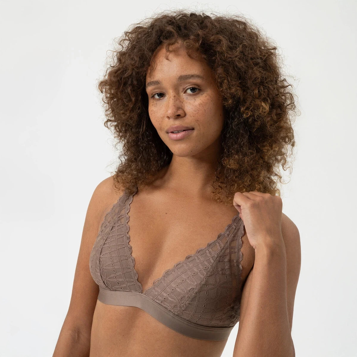 A woman with curly brown hair wearing a taupe lace bralette, posing against a clean white studio background.