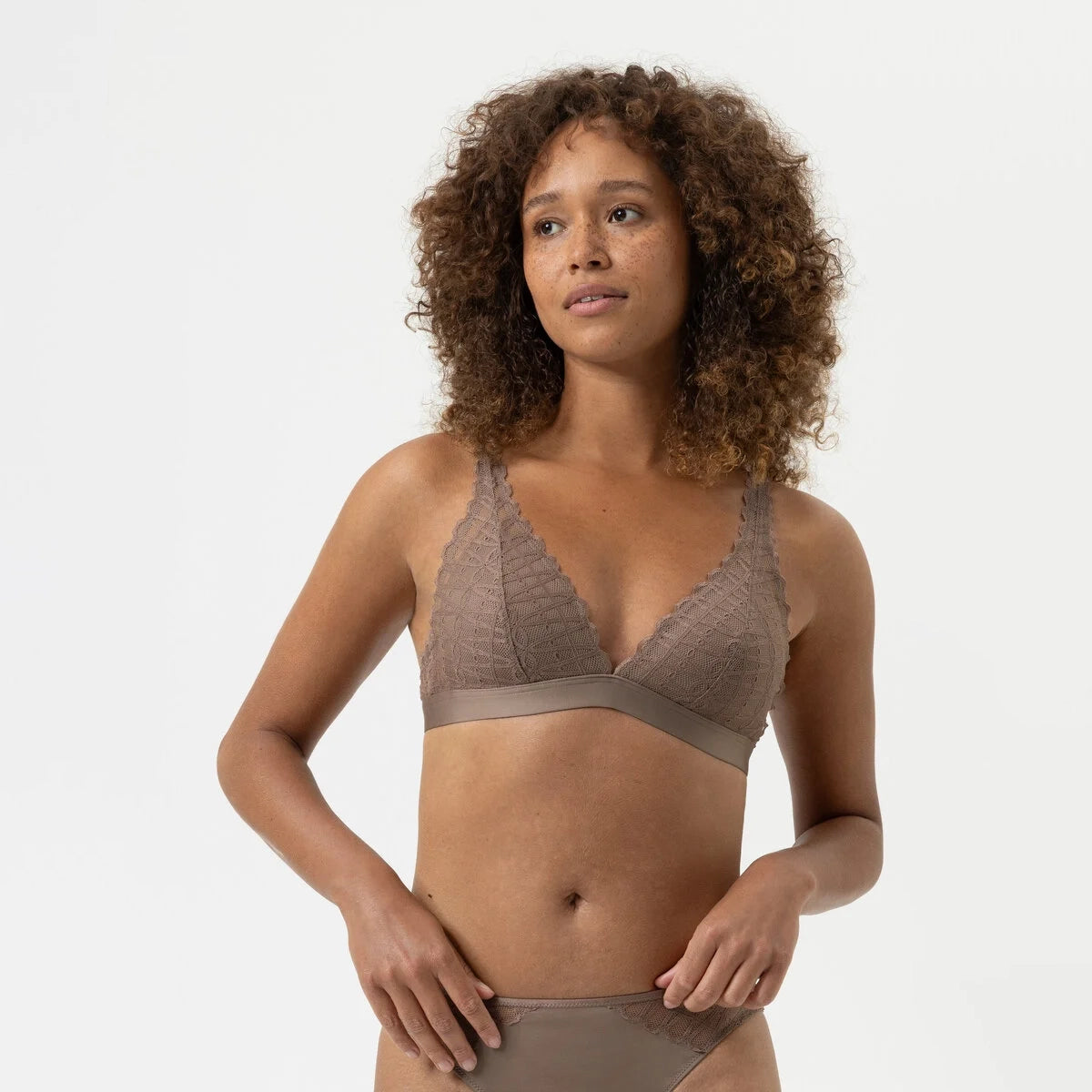 A woman with curly brown hair wearing a taupe lace bralette and matching underwear, photographed against a clean light gray studio background.