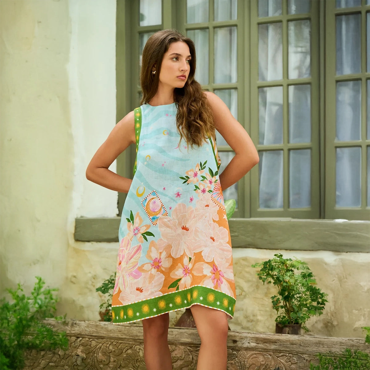 woman wearing a colourful floral dress standing in front of a window with green frames.