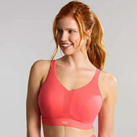 a smiling woman with red hair in a ponytail wearing a coral pink sports bra, photographed against a light gray studio background.