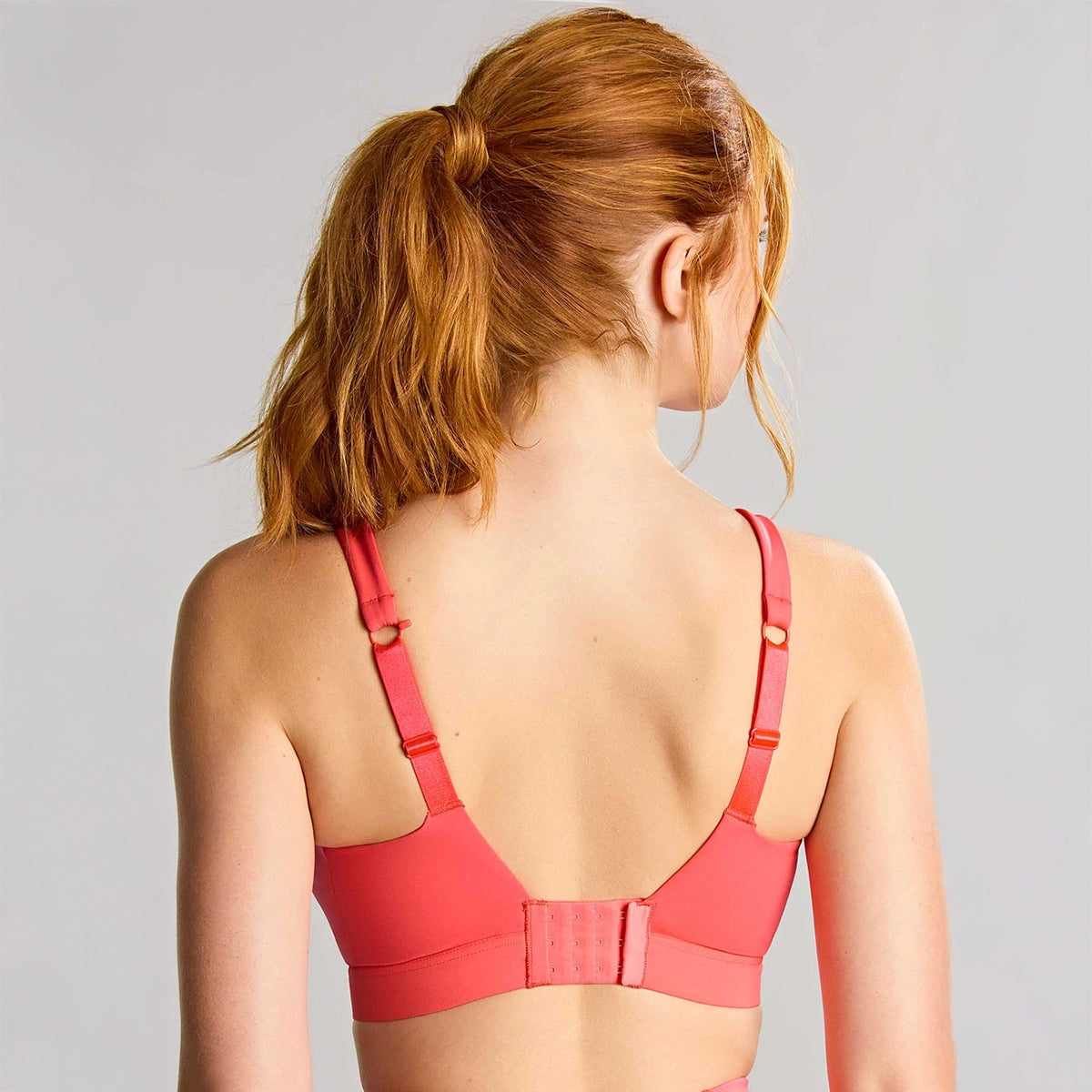 a woman with red hair tied back in a ponytail is shown from behind wearing a coral pink sports bra with adjustable straps and a hook-and-loop back closure, against a light gray studio background.