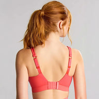 a woman with red hair tied back in a ponytail is shown from behind wearing a coral pink sports bra with adjustable straps and a hook-and-loop back closure, against a light gray studio background.