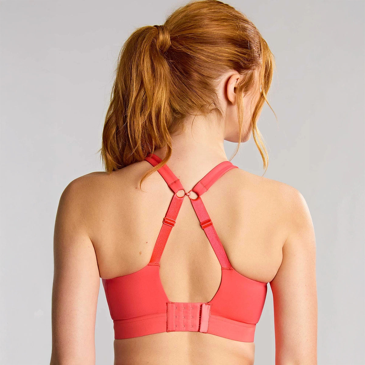 a woman with red hair in a ponytail is shown from the back wearing a coral pink sports bra with crisscrossing shoulder straps and a hook-and-loop closure at the lower back, set against a plain light gray studio background.