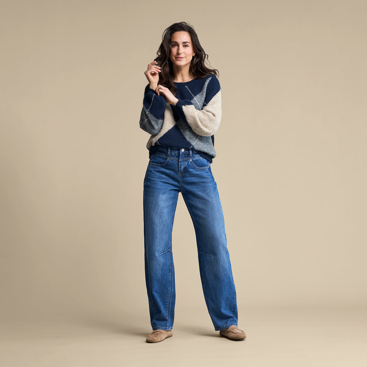 A woman modeling a navy and cream knit sweater with blue wide-leg jeans and tan shoes, standing against a solid beige studio background.