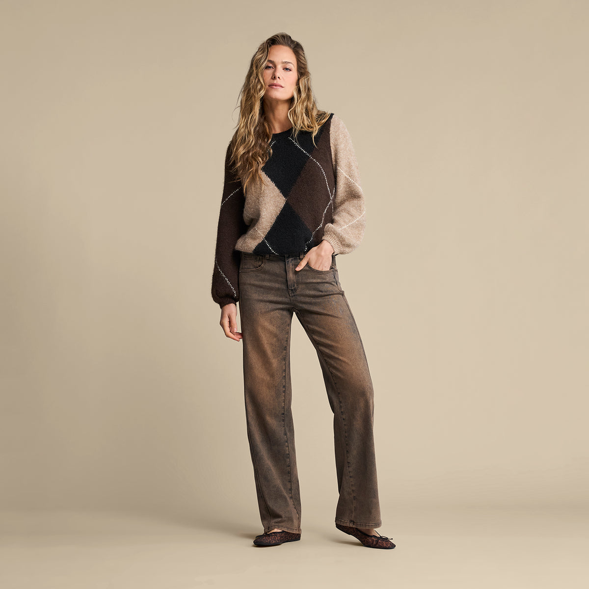 A woman stands against a plain beige studio background wearing a color-block knit sweater with dark brown, black, and beige panels, dark brown wide-leg jeans, and dark flat shoes.