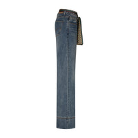 A pair of high-waisted blue denim jeans shown from the side, featuring a patterned scarf tie belt in beige, green, and black, set against a plain white background.