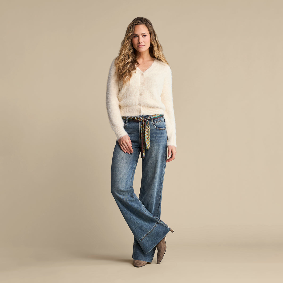A model wearing jumper and a pair of high-waisted blue denim jeans with a patterned scarf tie belt in beige, green, and black, set against a plain  background.