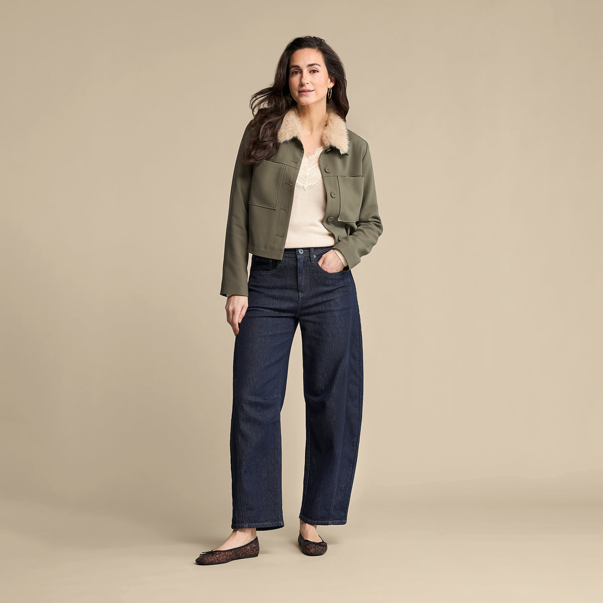 A woman stands against a plain beige studio background wearing an olive green cropped jacket with a light beige shearling-style collar over a cream top and dark blue wide-leg jeans.