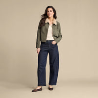 A woman stands against a plain beige studio background wearing an olive green cropped jacket with a light beige shearling-style collar over a cream top and dark blue wide-leg jeans.