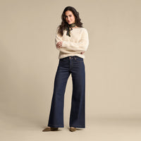 A woman modeling a cream knit sweater with a black neck scarf and dark blue wide-leg jeans, standing against a solid beige studio background.