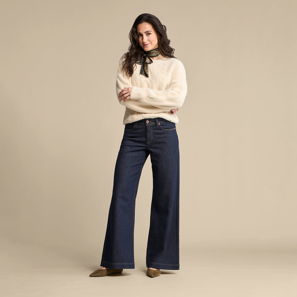 A woman modeling a cream knit sweater with a black neck scarf and dark blue wide-leg jeans, standing against a solid beige studio background.