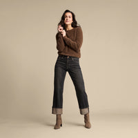 A woman modeling a brown knit sweater and dark blue cropped jeans with rolled cuffs, paired with brown ankle boots, against a solid beige studio background.