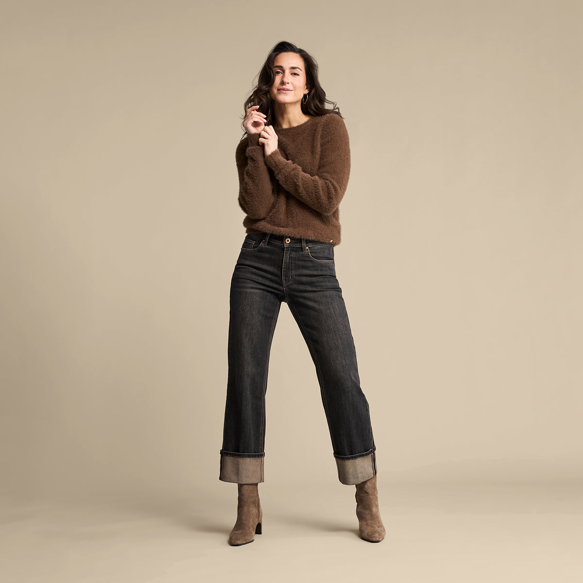A woman modeling a brown knit sweater and dark blue cropped jeans with rolled cuffs, paired with brown ankle boots, against a solid beige studio background.