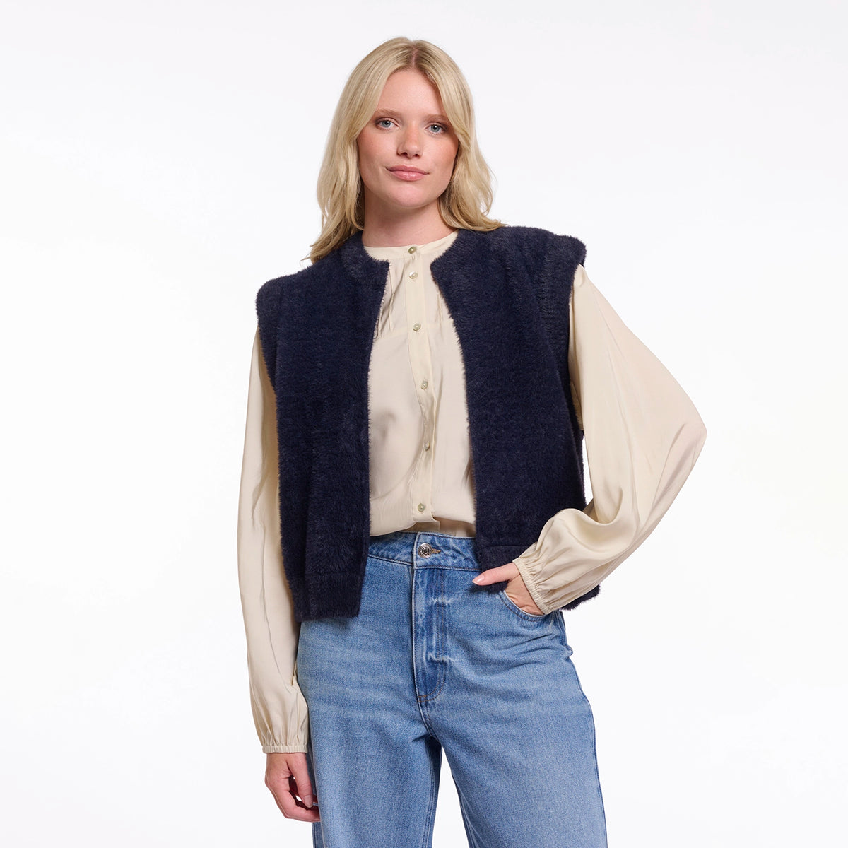 A blonde woman modeling a dark navy blue fuzzy sleeveless vest over a cream satin blouse and blue jeans, photographed against a clean white studio background.