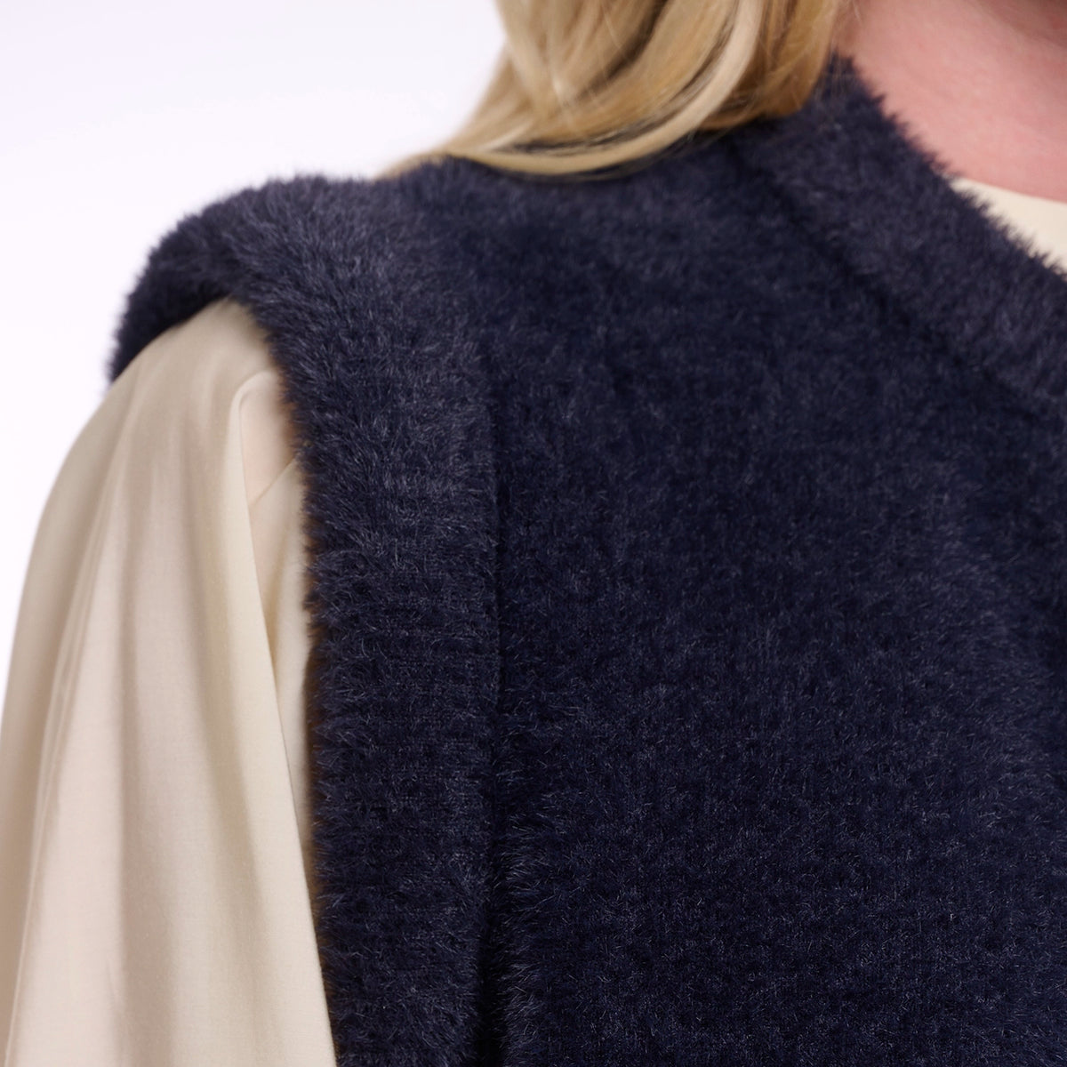 Close-up of a person wearing a dark navy fuzzy sleeveless vest over a cream-colored blouse, with blonde hair visible at the top.
