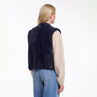 A woman shown from the back wearing a dark navy blue fuzzy sleeveless vest over a beige long-sleeve shirt and blue jeans, against a plain white studio background.