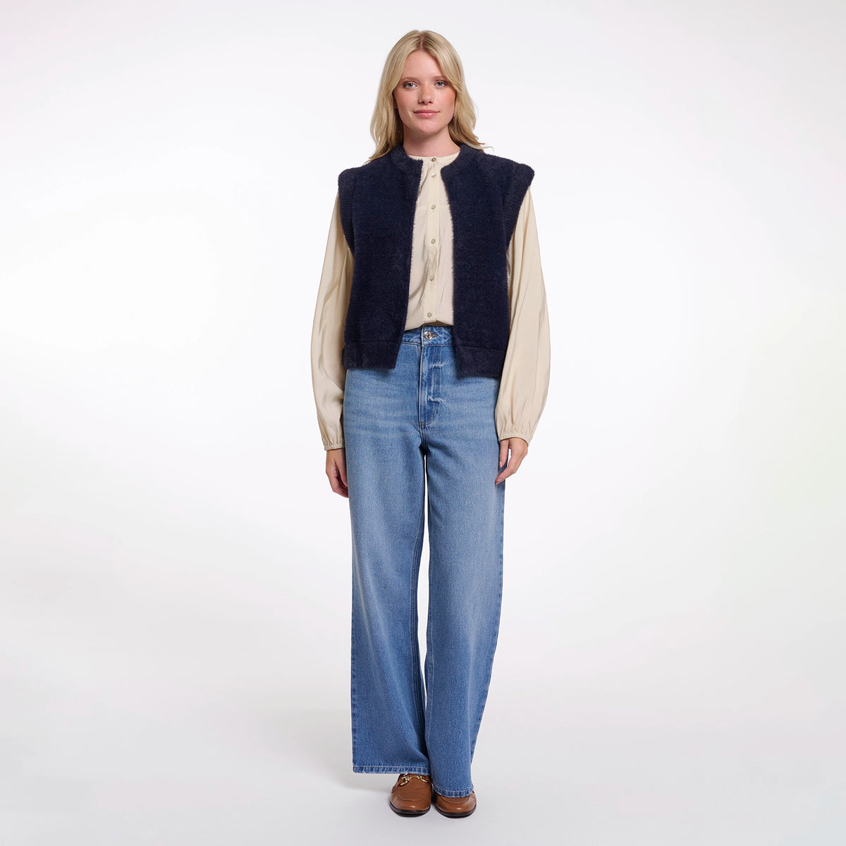 A woman stands against a plain white studio background wearing a dark navy sleeveless corduroy vest over a cream button-up shirt, paired with wide-leg blue jeans and brown shoes.