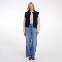 A woman stands against a plain white studio background wearing a dark navy sleeveless corduroy vest over a cream button-up shirt, paired with wide-leg blue jeans and brown shoes.
