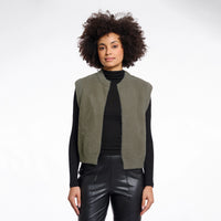 A model with curly dark hair is wearing a sleeveless, fuzzy olive green vest over a black turtleneck and black faux-leather pants, photographed against a solid white studio background.