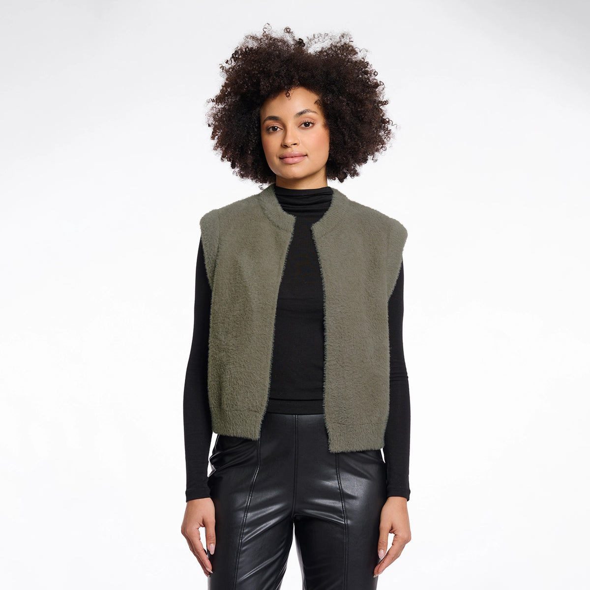 A model with curly dark hair is wearing a sleeveless, fuzzy olive green vest over a black turtleneck and black faux-leather pants, photographed against a solid white studio background.
