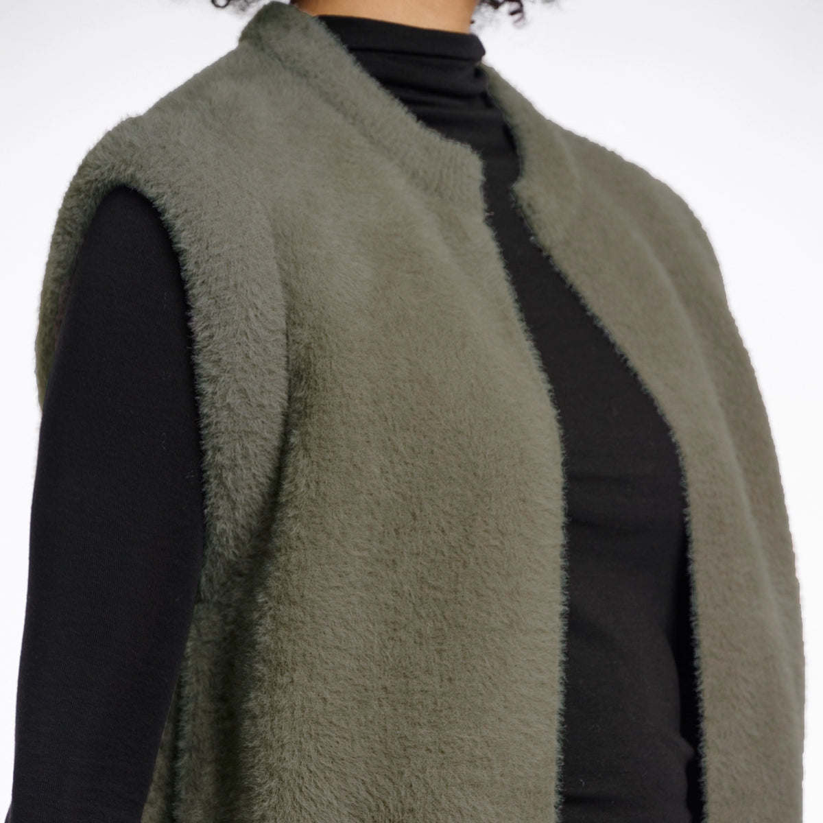An olive green fuzzy sleeveless vest layered over a black long-sleeve top, shown on a model against a plain white background.