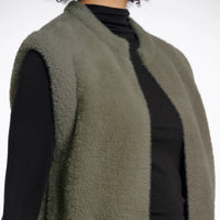 An olive green fuzzy sleeveless vest layered over a black long-sleeve top, shown on a model against a plain white background.