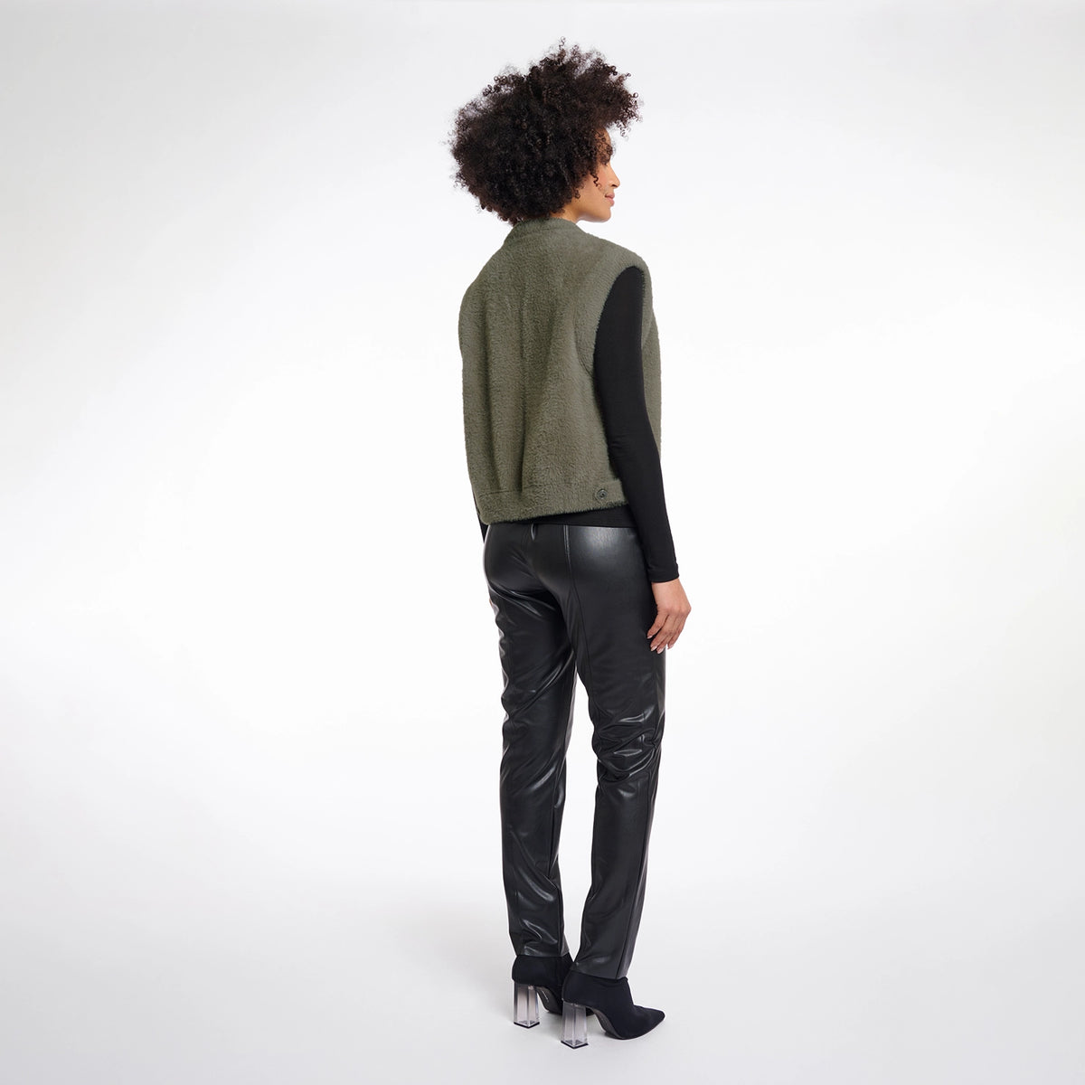 A model shown from the back wearing an olive green knit sleeveless vest over a black long-sleeve top, paired with black faux-leather pants and black high-heeled boots, against a white studio background.