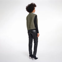 A model shown from the back wearing an olive green knit sleeveless vest over a black long-sleeve top, paired with black faux-leather pants and black high-heeled boots, against a white studio background.