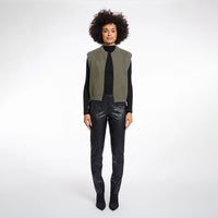 A model with an afro hairstyle stands against a light gray studio background wearing a sleeveless vest with olive green and black panels over a black long-sleeve top and black faux-leather pants.