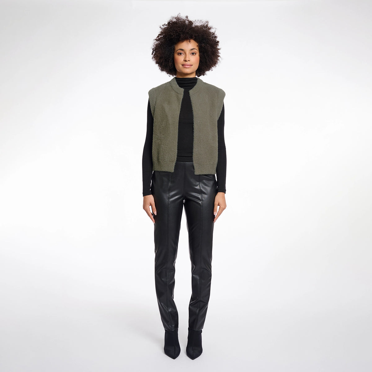 A model with an afro hairstyle stands against a light gray studio background wearing a sleeveless vest with olive green and black panels over a black long-sleeve top and black faux-leather pants.