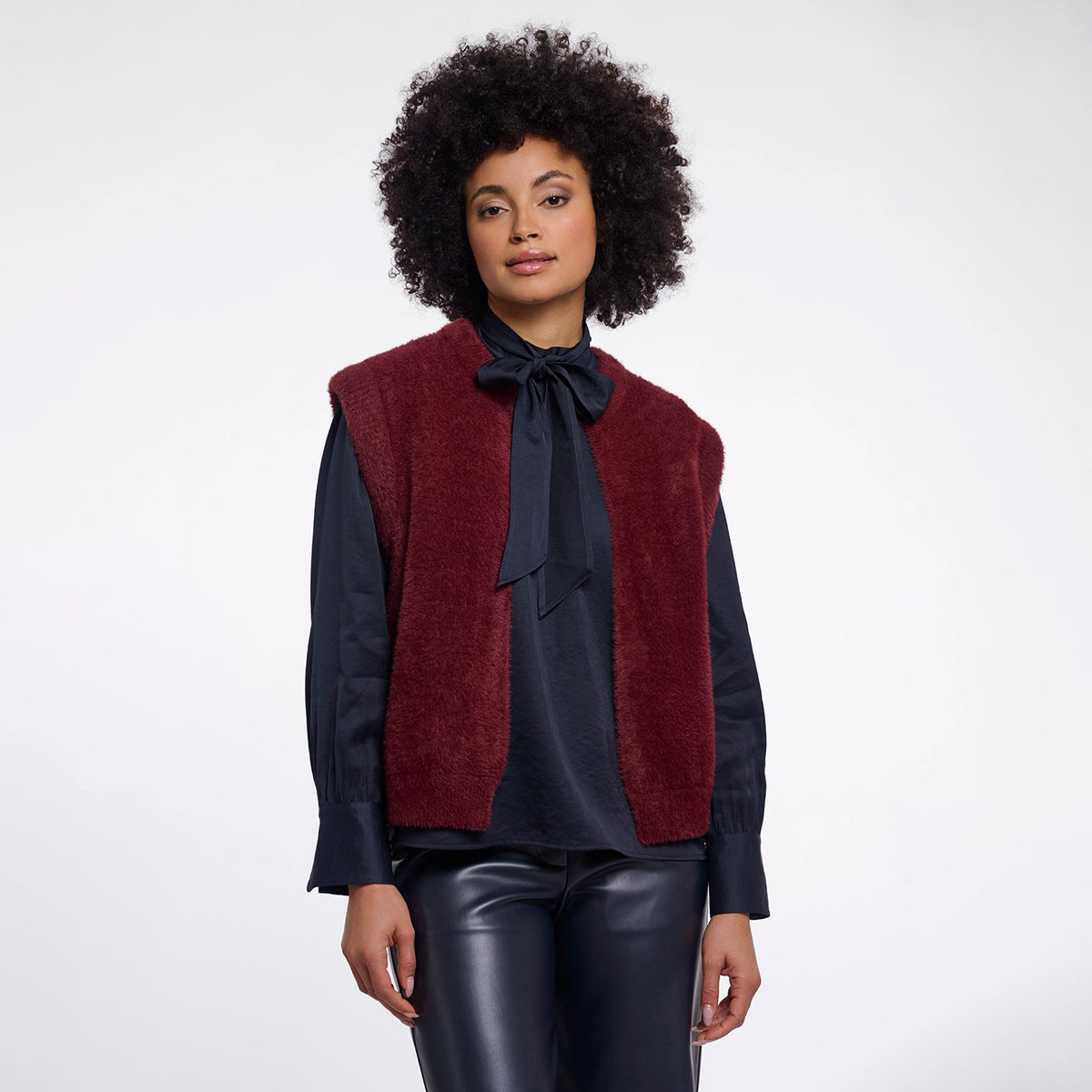 A model with curly black hair is wearing a burgundy fuzzy knit vest over a navy blue long-sleeve blouse with a large bow, paired with dark shiny pants, against a plain light gray background.