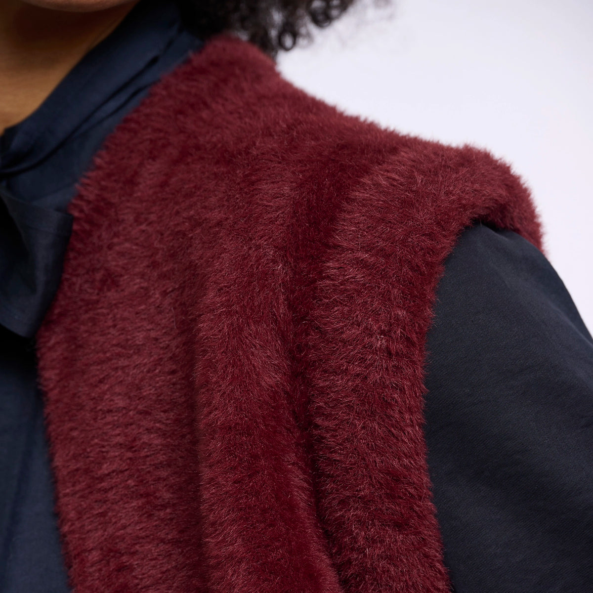 A close-up of a person wearing a plush, shaggy burgundy faux-fur vest over a dark navy shirt against a plain light background.