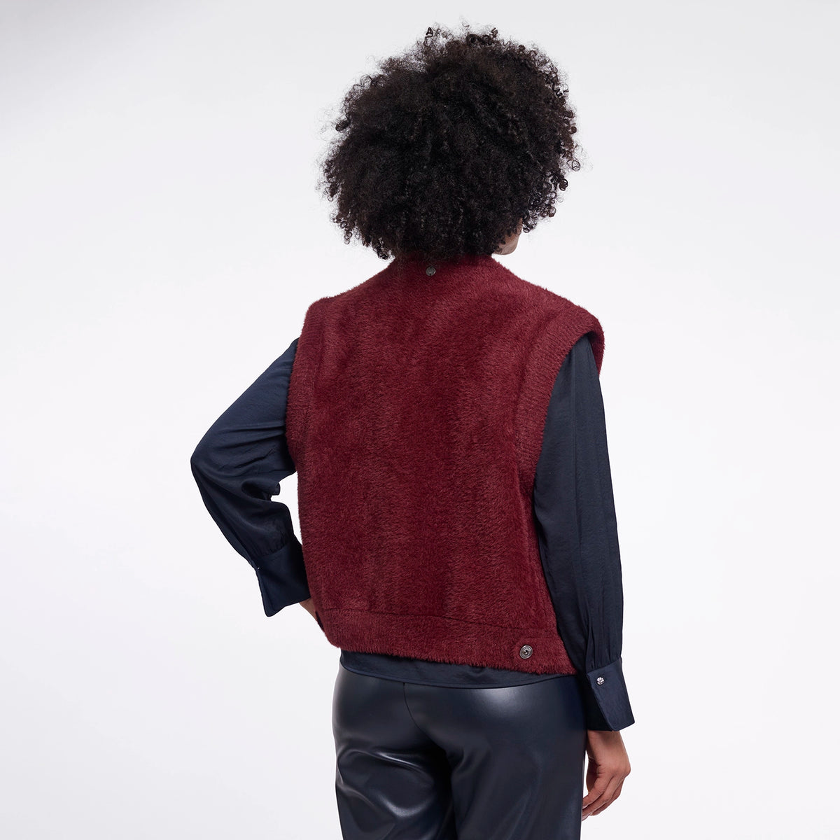 A person shown from the back wearing a sleeveless burgundy sherpa vest over a dark navy long-sleeve shirt and black pants, photographed against a clean white studio background.