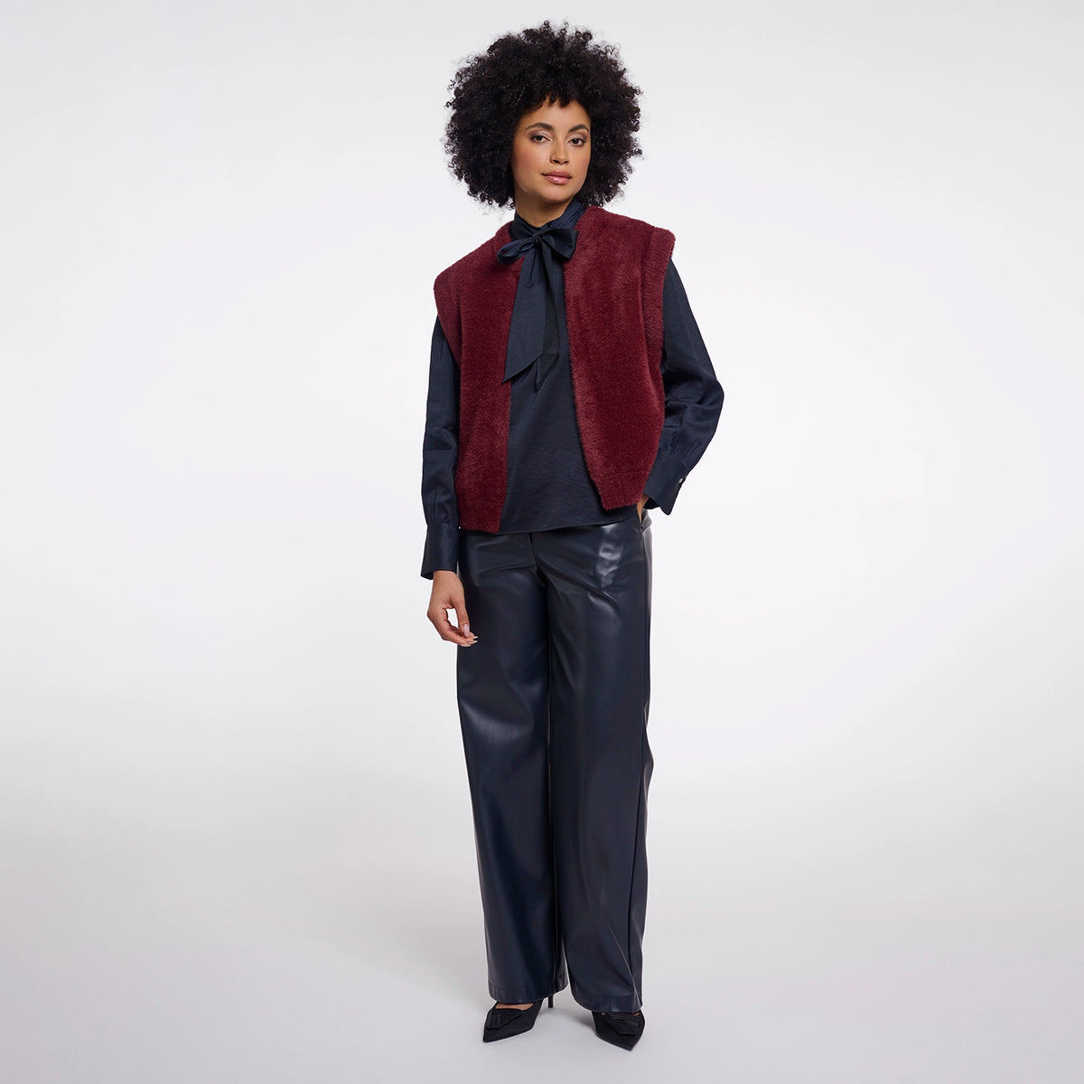 A model with an afro hairstyle stands against a light gray studio background wearing a burgundy faux-fur vest over a dark navy long-sleeve shirt with a bow tie, paired with dark navy wide-leg trousers and black pointed shoes.