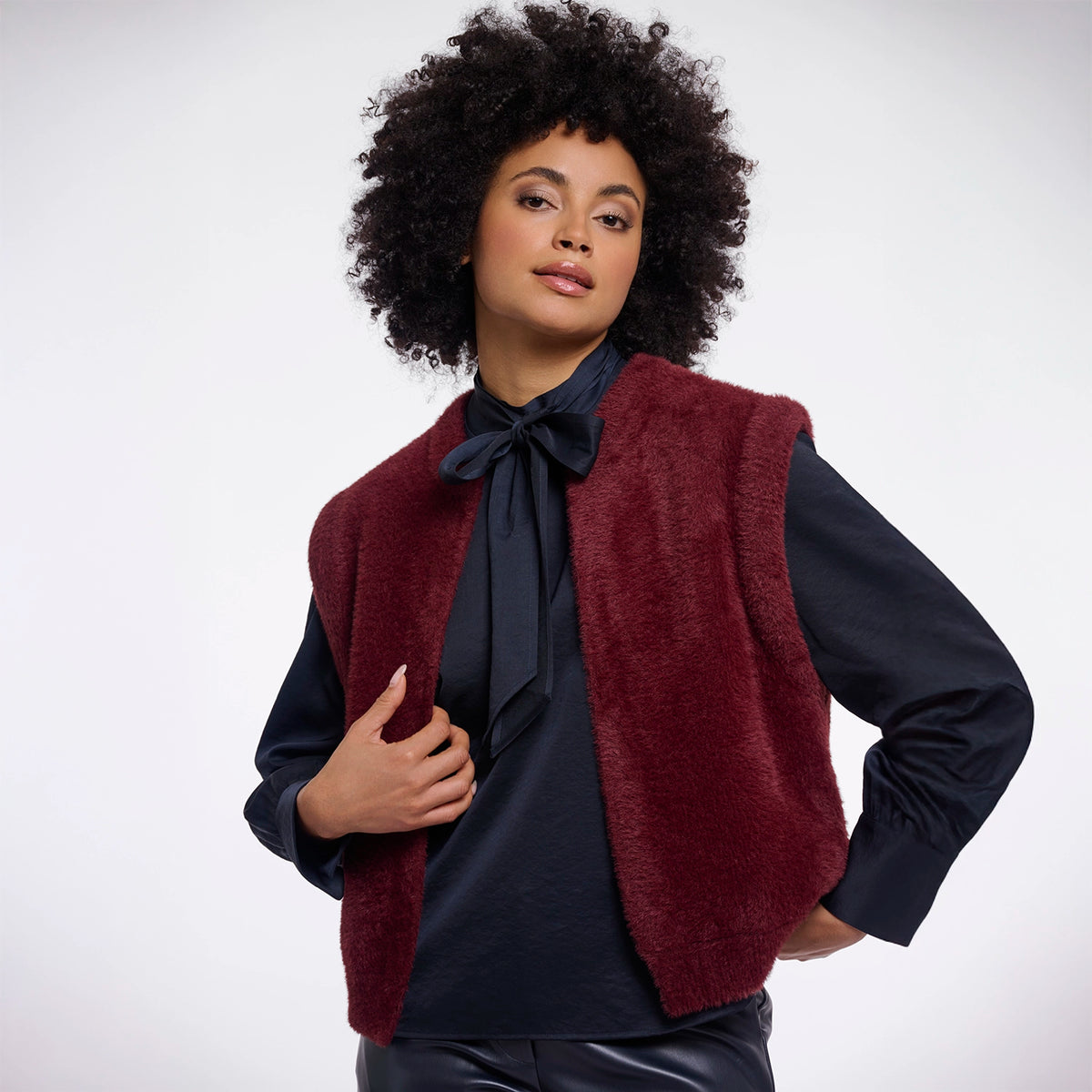 A model with an afro hairstyle wearing a burgundy fuzzy sleeveless vest over a dark navy satin blouse with a large bow at the collar, paired with dark leather pants, against a light gray studio background.