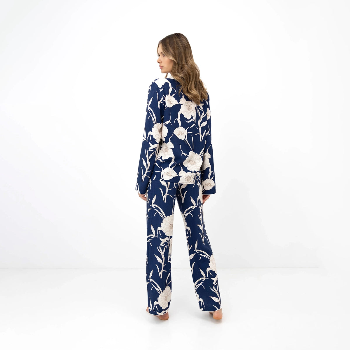A woman stands with her back to the camera wearing a matching two-piece pyjama set with a navy blue base and a large white floral print, set against a plain white studio background.