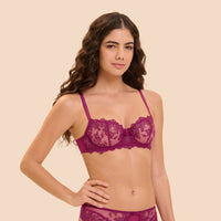 A woman modeling a deep magenta lace bra and matching panties against a solid beige background.