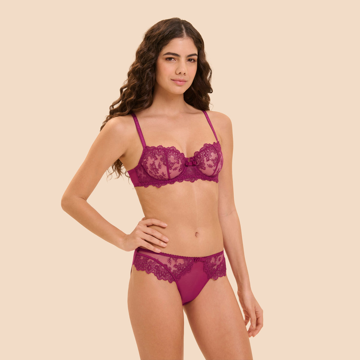 A woman modeling a matching magenta lace lingerie set, including a sheer underwire bra and coordinating panties, against a solid light beige background.