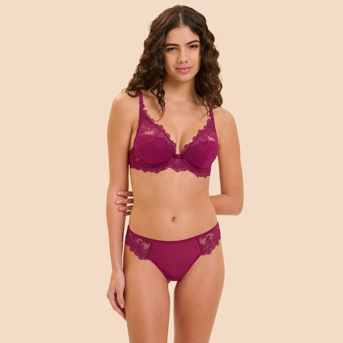 A woman modeling a matching lingerie set in a deep magenta color, featuring a lace underwire bra and coordinating lace-trimmed panties, set against a solid beige background.