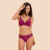 A woman modeling a matching magenta lace bra and panty set against a solid beige background.