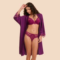 A woman modeling a matching lingerie set with a sheer purple robe. The set includes a lace bra and panties in a deep purple shade, with the robe featuring matching purple lace trim on the sleeves and hem, set against a solid beige background.