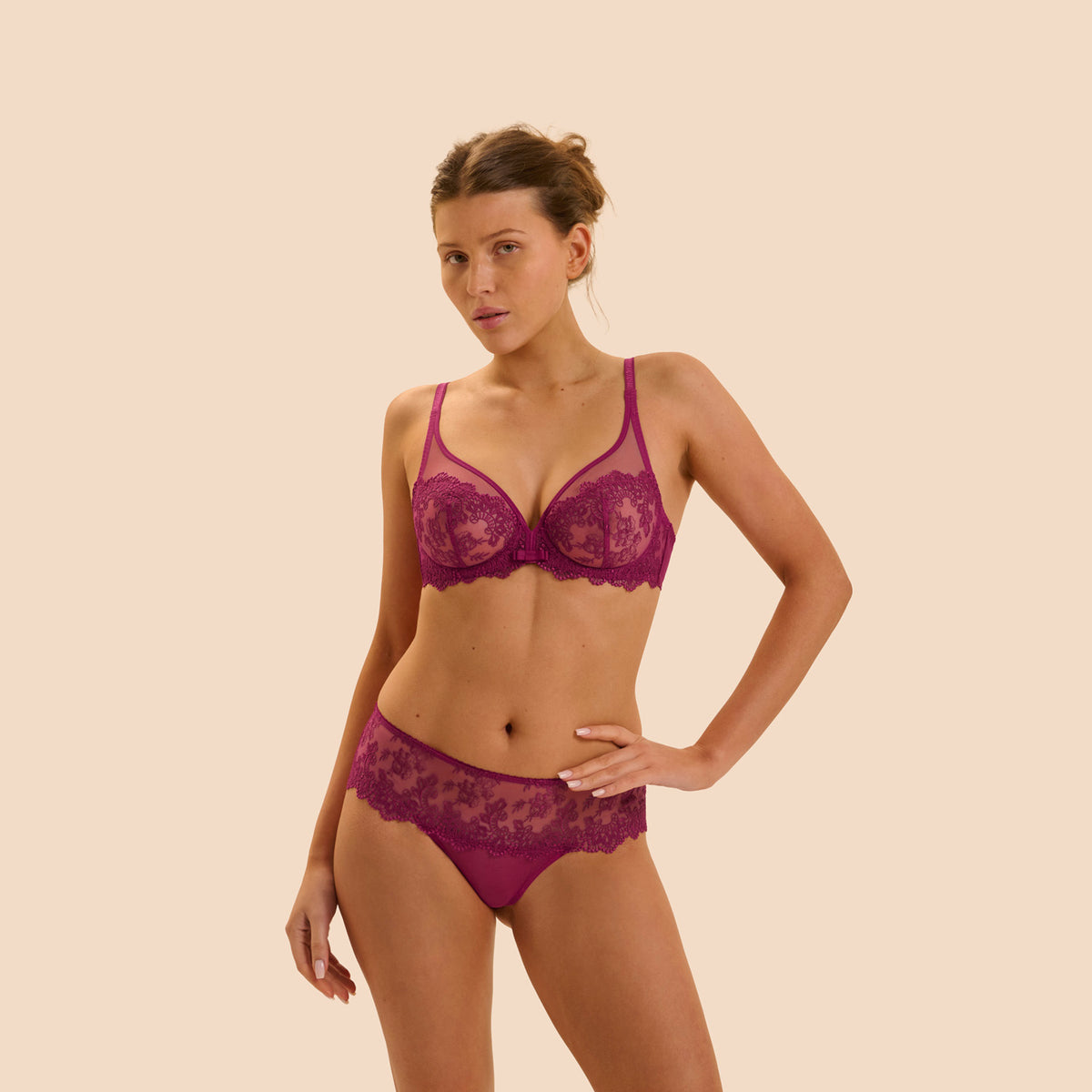 A woman modeling a matching magenta lace bra and panty set against a solid beige background.