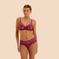 A woman modeling a matching lace lingerie set in a deep magenta-purple color, consisting of a sheer underwire bra and coordinating panties, set against a solid pale beige background.