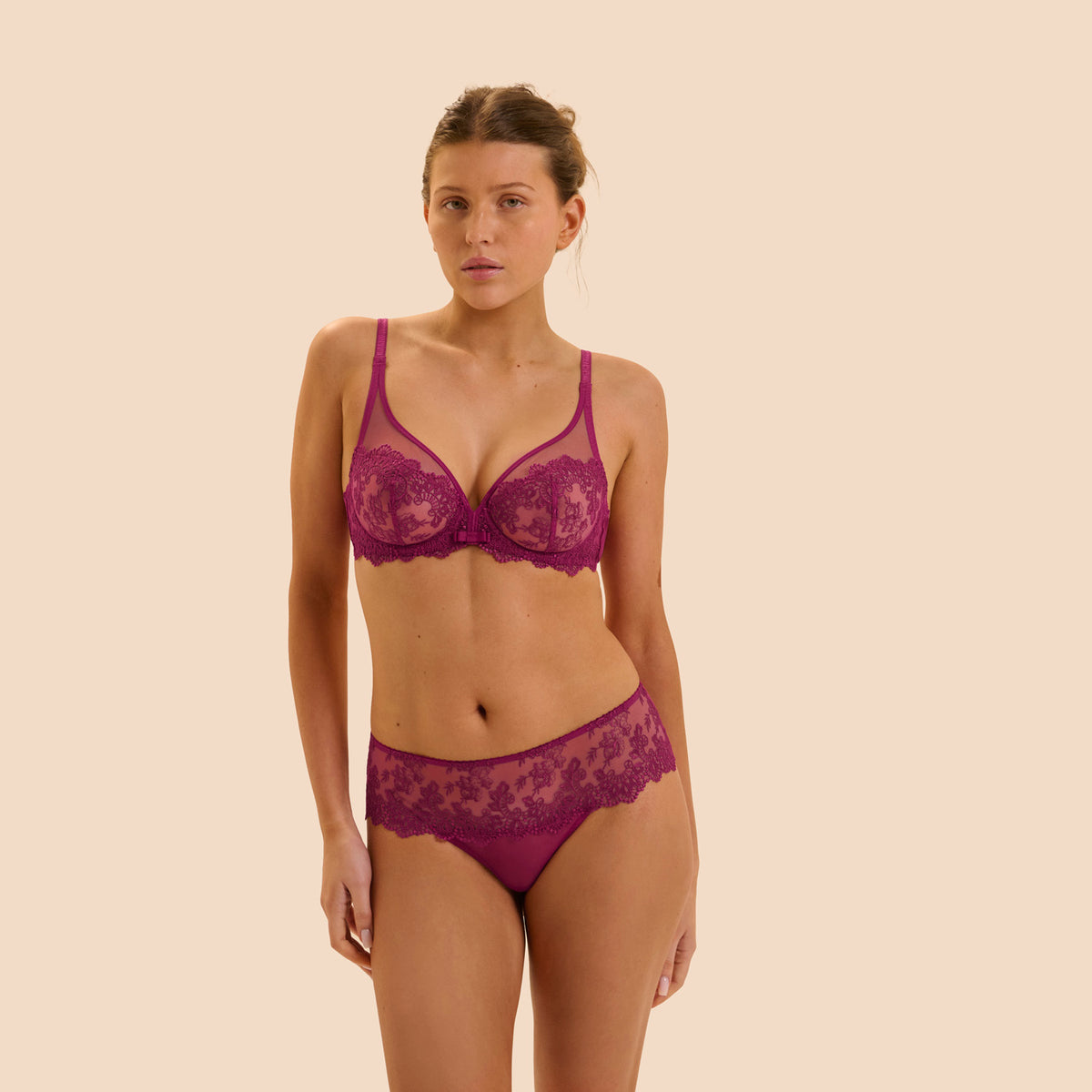 A woman modeling a matching lace lingerie set in a deep magenta-purple color, consisting of a sheer underwire bra and coordinating panties, set against a solid pale beige background.
