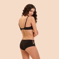 A woman modeling black lingerie, shown from the back, wearing a black bralette and matching high-waisted briefs with lace detailing on the side, set against a solid beige background.