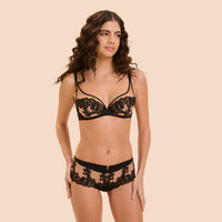 A woman modeling a black lace lingerie set against a solid beige background. The set includes a sheer underwire bra with floral lace cups and matching high-waisted briefs with a lace waistband.
