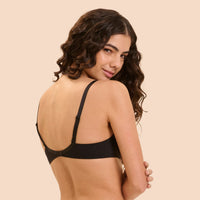 A woman with long, wavy dark brown hair is shown from the back wearing a black bralette against a solid light beige background.