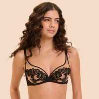 A woman modeling a black lace underwire bra with sheer mesh cups against a solid beige background.
